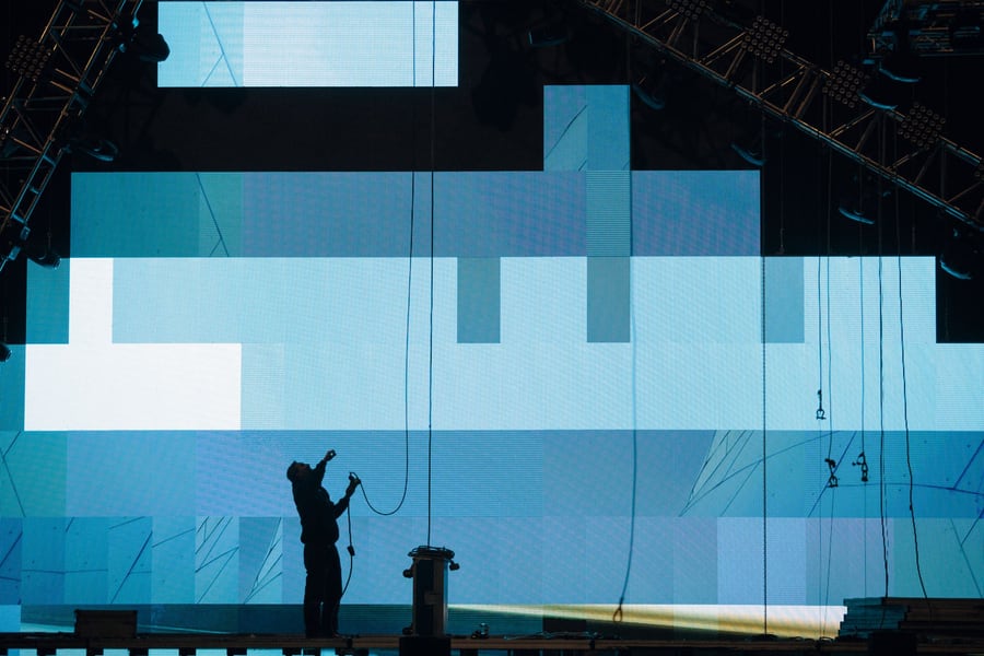 A vast pale-blue LED wall with a rigger silhouetted at its base